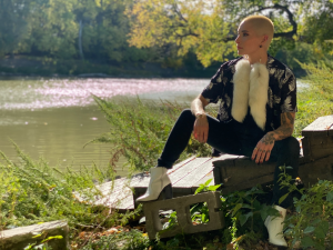 Adrienne kneels next to a river. They are wearing a short-sleeved collared shirt, with black pants, white boots, and long, white, furry earrings. 