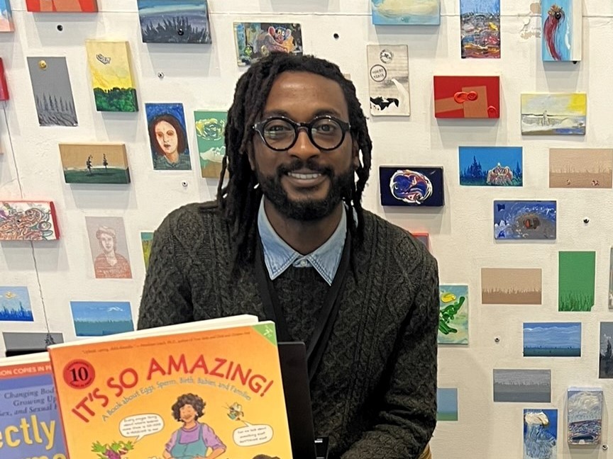 Hafiz Jatto sitting in front of a wall covered in many tiny paintings. In front of Hafiz is the book "It's So Amazing!".